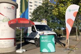 Infostand Digtialer Engel mit Litfaßsäule, Sonnenschirm, Tresen. Infomobil im Hintergrund.