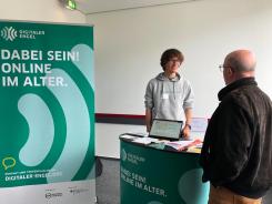 Mobilreferent Johannes ist im Gespräch mit einem Fachbesucher am Messestand des Digitalen Engel. Auf der linken Seite steht ein Roll Up mit der Aufschrift "Dabei sein! Online im Alter."