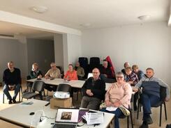 Zwöf Teilnehmende sitzen auf Stühlen und blicken nach vorne. Am vorderen Bildrand sieht man einen Laptop und einen Beamer.