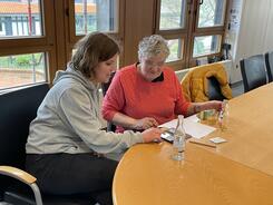 Theresa sitzt mit einer Besucherin am Tisch und erklärt Dinge am Smartphone.