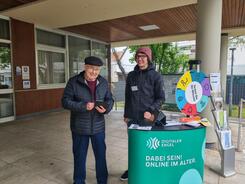 Johannes steht hinter dem Infostand und blickt zu einem Herrn der neben ihm steht und auf sein Smartphone schaut