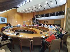 Das Foto im Weitwinkelmodus zeigt den Sitzungssaal des Rathauses. In einem Kreis angeordnet sitzen ca. 25 Senior:innen. Viele gucken in die Kamera.