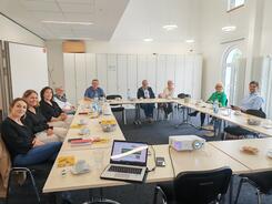Mehrere Personen sitzen an einem großen zusammengeschobenen Tisch im Raum und blicken nach Vorne 