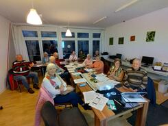In einem Raum im MGH Buntes Haus sitzen vier ältere Männer und acht ältere Frauen an einem länglichen Tisch. Die meisten blicken nach vorne in die Kamera. Auf dem Tisch im vorderen Bildrand stehen ein Beamer und ein Laptop.