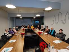 14 Seniorinnen und Senioren sitzen in einem großen Seminarraum im Stadtarchiv an einem langen Tisch in U-Form. Sie gucken nach vorne in die Kamera. Auf dem Tisch liegen Winterdekorationen und Unterlagen vom Digitalen Engel.