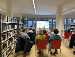Die Teilnehmenden sitzen in einer Bibliothek auf roten Stühlen und sind von hinten zu sehen. Sie schauen auf die Titelfolie zum Thema hilfreiche Apps im Alltag älterer Menschen.