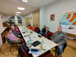 An einem langen Tisch sitzen dreizehn Senior:innen und blicken nach vorne in die Kamera. Am Bildrand rechts hängt ein Plakat vom Digitalen Engel mit Informationen zu der Veranstaltung.