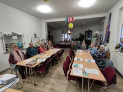 An zwei Tischen sitzen sieben Senior:innen und blicken nach vorne. Auf den Tischen liegen Materialien vom Digitalen Engel,