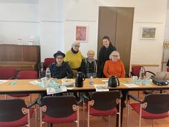 3 Teilnehmende sitzen auf schwarz-roten Stühlen, zwei Teilnehmerinnen stehen hinter ihnen. 