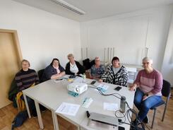 Sechs Frauen sitzen an zwei zusammengestellten Tischen und gucken in die Kamera. Auf den Tischen liegen ein Beamer, ein Laptop und Unterlagen.