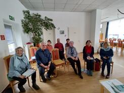 Acht Senior:innen (zwei Männer und sechs Frauen) sitzen in einem Raum auf Stühlen und lächeln nach vorne in die Kamera.