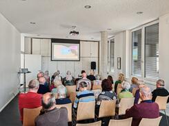 Mehrere Personen sitzen auf Stühlen im Raum und blicken nach Vorne auf eine Leinwand auf der ein Video abgespielt wird.