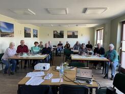 Die 13 Teilnehmenden sitzen an Tischen mit bis zu vier Personen und lächeln in die Kamera.