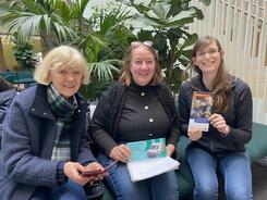 Petra (rechts) sitzt mit den beiden Freundinnen Astrid (Mitte) und Christine (links) im Atrium des KörberHauses. Petra und Astrid halten zwei Flyer in den Händen und Christine ihr Smartphone.