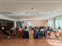 Zahlreiche Personen sitzen auf Stühlen im Raum verteilt und blicken nach Vorne zu einer Leinwand.
