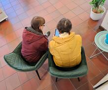 Petra (links) sitzt neben einer Seniorin mit einer gelben Jacke. Die beiden schauen sich gemeinsam ein Smartphone an. Das Bild ist aus der Vogelperspektive entstanden.