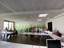 In einem großen Raum sitzen insgesamt 15 Menschen. Das Foto ist im Weitwinkel-Modus aufgenommen, damit alle Teilnehmende, die im Raum verteilt sitzen, abgebildet sind.