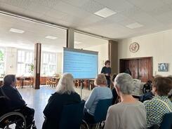 Mehrere Personen sitzen auf Stühlen im Raum und blicken nach Vorne zu einer Leinwand und zu Referent Johannes.