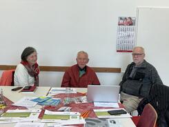 Die drei Teilnehmenden sitzen an einem mit Broschüren und Handouts gedeckten Tisch und lächeln in die Kamera.