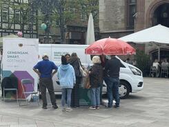 Aus der Ferne ist der Info-Stand mit dem Info-Mobil fotografiert. Es stehen mehrere Passant:innen an dem Stand und sind im Gespräch. 