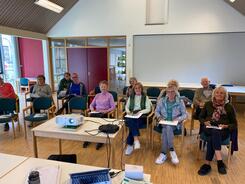 Im Dorftreff Ankum sitzen in einem Raum auf Stühlen elf Personen und blicken in die Kamera. Vor ihnen steht ein Tisch mit einem Beamer.