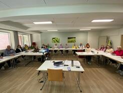 Ein Schulungsraum mit einer Gruppe älterer Menschen, die in einem U-förmig angeordneten Sitzkreis an Tischen sitzen. Alle Teilnehmenden schauen freundlich in die Kamera. Auf den Tischen liegen Unterlagen, Stifte und digitale Geräte. Im Vordergrund ist ein Laptop und ein Beamer zu sehen.
