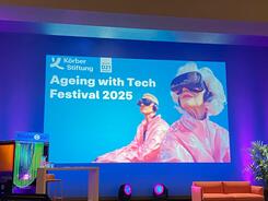 Eine Bühne im Theatersaal des KörberHauses. Eine Präsentation zeigt eine Folie mit der Überschrift "Ageing with Tech Festival 2025". Rechts ist eine KI-generiertes Bild mit einer älteren Dama und einer jüngeren Person, die eine VR-Brille tragen.