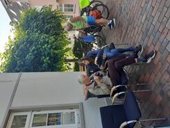 Auf dem Bild im Hochkantformat sitzt Petra mit einem älteren Herr auf zwei Stühlen. Sie erklärt ihm etwas auf dem Smartphone. Rechts im Hintergrund steht ein sommerlich gekleideter Mann an einem Fahrrad.