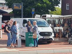 Petra steht mit einer Seniorin am Info-Stand vor dem Info-Mobil. Das Bild ist aus der Ferne aufgenommen. Links von den beiden stehen zwei weitere Frauen. Rechts ist ein Stand mit Taschen.