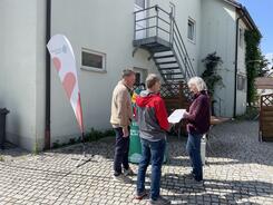 Zwei Besucher und eine Besucherin stehen an unserem Infostand und von hinten zu sehen. Sie unterhalten sich.