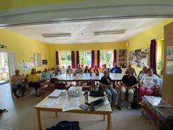 Im Gemeindehaus in Ahrensbök mit gelben Wänden sitzen an einem landen ovalen Tisch 15 Senior:innen, davon drei Männer. Im vorderen Teil steht ein Tisch mit einem Beamer drauf.