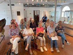 In einem Museumraum sitzen knapp fünfzehn Senior:innen in drei Stuhlreihen und schauen nach vorne in die Kamera.
