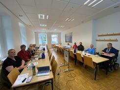 In einem länglichen Raum sitzen an zwei langen Tisch knapp 12 Personen. Auf den Tischen liegen Materialien und Getränke. Zwischen den Tischen steht ein Beamer auf einer Erhöhung.