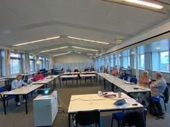 In einem sehr großen Raum sitzen ca. 13 Personen an Tischen, die in einer U-Form aufgestellt sind. Im Vordergrund steht ein Tisch mit einem Laptop und ein kleiner Schrank mit einem Beamer.