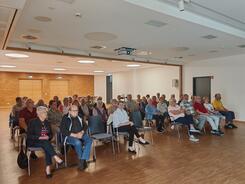 Zahlreiche Personen sitzen auf Stühlen in einem großen Saal und blicken nach Vorne.