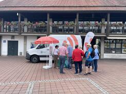 Das Infomobil inklusive des Standes steht vor einer Markthalle auf einem Marktplatz. Aufgrund des Wetters steht ein Sonnenschirm am Stand. Eine Gruppe von Senior:innen steht am Infostand.