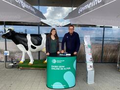 Petra (links) steht mit Bürgermeister Jürgen Schiewer (rechts) am Info-Stand vor einem Supermarkt. Links neben Petra steht eine lebensgroße Kuh-Figur.