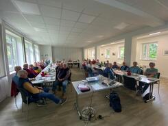 Das Foto ist im Weitwinkelmodus aufgenommen. Es zeigt über dreißig Personen, die an Tischen in U-Form sitzen. Im Vordergrund steht ein Tisch mit einem Laptop und Beamer.