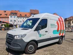 Das Infomobil steht auf einem gepflasterten Platz mit Häusern im Hintergrund.