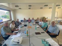 Elf Senior:innen sitzen im Dörphus an einem großen, rechteckigen Tisch. Sie blicken nach vorne in die Kamera. Auf dem Tisch liegen Unterlagen, Wasserflaschen und ein Beamer.