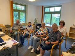 Ein Mann und vier Frauen sitzen nebeneinander auf Stühlen und blicken zur Seite in die Kamera.