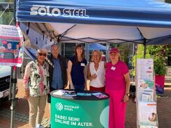 Petra (Mitte) posiert mit vier Senior:innen (drei Frauen, ein Mann) am Info-Stand unter einem Pavillon in die Kamera.