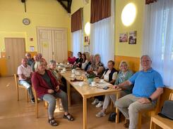 In der DRK-Sozialstation sitzen zehn Senior:innen und die drei Schülerinnen an einem langen Tisch und gucken nach vorne in die Kamera. Auf dem Tisch liegen Materialien und Kaffeegedeck.