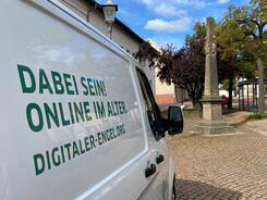 Links im Vordergrund ist das Info-Mobil mit folgender Aufschrift abgebildet: "Dabei sein - Online im Alter! digitaler-engel.org". Rechts im Hintergrund steht die Postsäule.
