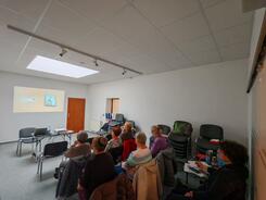 Eine Gruppe von Menschen sitzt in einem Raum und blickt nach Vorne zu einer Wand auf die ein Video projiziert wird.