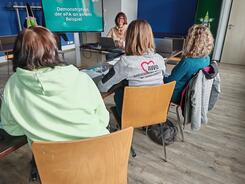 Drei Frauen sitzen auf Stühlen an einem Tisch und blicken nach Vorne auf einen Bildschirm und zum Referenten, der am Laptop sitzt.