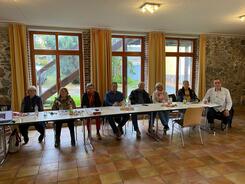 An einem langen Tisch sitzen acht Personen (drei Männer und fünf Frauen) und gucken nach vorne in die Kamera.