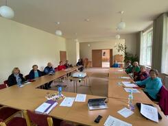 Im Dorfgemeinschaftshaus sitzen rund zehn Senior:innen links und rechts im Raum an Tischen und blicken nach vorne. Auf den Tischen, die in U-Form gegliedert sind, liegen Materialien, unter anderem auch der Laptop von Petra. In der Mitte des U steht ein Beamer auf einem Tisch.