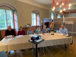 In einem Raum sitzen an einem Tisch sechs Senior:innen (vier Frauen in der Mitte und zwei Männer an den Rändern). In der Mitte steht ein Beamer.