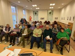 In einem gelb erleuchteten Raum sitzen in vier Sitzreihen 25 Senior:innen. Sie blicken nach vorne in die Kamera.